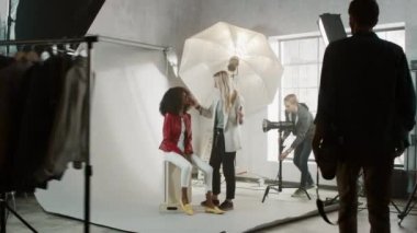 Fotoğraf çekiminin sahne arkası: Makyöz, gür kıvırcık saçlı güzel siyah modele ruj sürüyor. Fotoğrafçı fotoğraf çekmeye hazır. Moda Dergisi Stüdyo Fotoğrafları