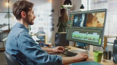 Beard ve Jeans Shirt Works 'ün erkek video editörü Big Explay ile Kişisel Bilgisayarında Görüntü. Havalı bir ofisin çatı katında çalışıyor. Diğer Yaratıcı Kadın Meslektaşlar Arkaplanda Yürüyor.