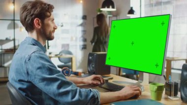Sakallı ve Kot Gömlekli Erkek Video Düzenleyicisi Büyük Yeşil Ekran Mock Up ile Kişisel Bilgisayarında çalışıyor. Havalı bir ofisin çatı katında çalışıyor. Diğer Yaratıcı Kadın Meslektaşlar Yürüyüşü