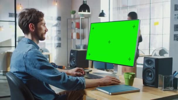 Manlig videoredigerare med skägg och Jeans tröja Fungerar på hans persondator med Big Green Screen Mock Up Display. Han jobbar på ett coolt kontor. Andra kvinnliga kreativa Colleague promenader i — Stockvideo