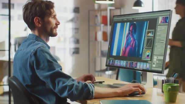 Man Digital Editor i Jeans Shirt Fungerar i fotoredigeringsprogram på sin personliga dator med stor skärm. Han jobbar på Cool Office Loft. Andra kvinnliga kreativa Colleague promenader i bakgrunden. — Stockvideo