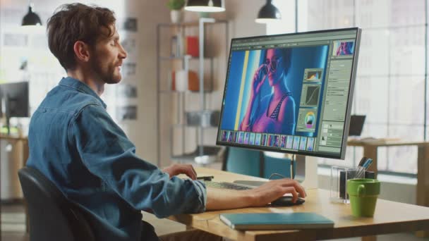 Мужчина в джинсах работает над редактированием изображений на своем персональном компьютере с большим экраном. Он работает в клёвом офисе Лофт. Другие женщины творческий коллега прогулки в фоновом режиме. — стоковое видео