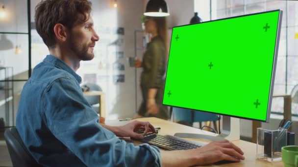 Manlig kreativ designer med skägg och Jeans tröja Fungerar på sin personliga dator med Big Green Screen Mock Up Display. Han jobbar på ett coolt kontor. Andra kvinnliga kreativa Colleague promenader i — Stockvideo