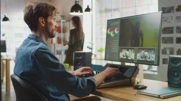 Editor de Vídeo Masculino com Barba e Camisa Jeans Funciona com Filmagem em Seu Computador Pessoal Usando um Console Deck de Edição. Ele trabalha em um escritório legal Loft. Outros Caminhadas Colega Criativa Feminino no — Vídeo de Stock