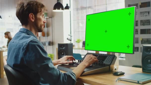 Young Professional Creative Employee funciona em seu computador pessoal com tela verde grande Mock Up Display. Ele trabalha em um escritório legal Loft. Ele está usando uma plataforma de edição de console em vez de teclado. — Vídeo de Stock