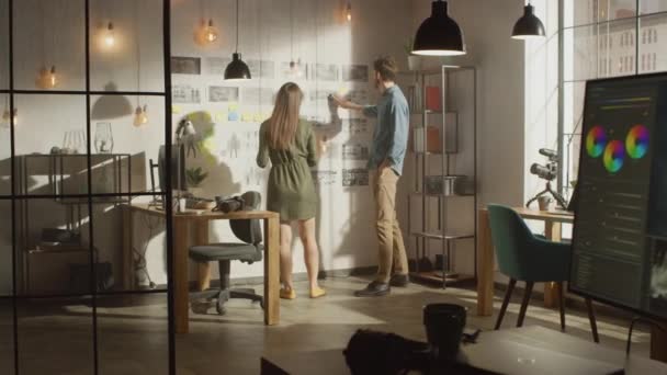 De jonge creatieve man en vrouw zijn het organiseren van een Mood Board op een muur van hun Cool Office Loft. Foto 's vertegenwoordigen Toekomstige Beweging Video. Collega 's werken en discussiëren over het project. Zonnige dag. — Stockvideo