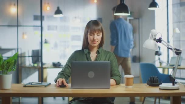 Joven hermosa morena trabaja en un ordenador portátil en Cool Creative Agency en una oficina Loft. Ella tiene un café para llevar y un cuaderno en su mesa. Su colega camina en el fondo. — Vídeos de Stock