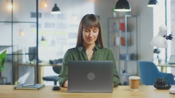 若い美しいブルネットはロフトオフィスのクールなクリエイティブエージェンシーのラップトップコンピュータで動作します。彼女はテイクアウェイコーヒーと彼女のテーブルのノートを持っています.カメラがズームインして笑顔で. — ストック動画
