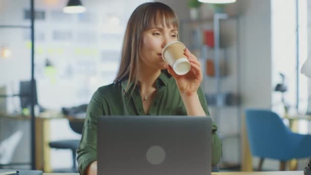 Young Beautiful Brunette Works on a Laptop Computer in Cool Creative Agency in a Loft Office Вона п'є каву з відніманням. Вихід з камери. — стокове відео