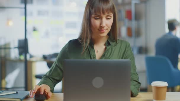 若い美しいブルネットはロフトオフィスのクールなクリエイティブエージェンシーのラップトップコンピュータで動作します。彼女はテイクアウェイコーヒーと彼女のテーブルのノートを持っています.カメラのズームアウト. — ストック動画