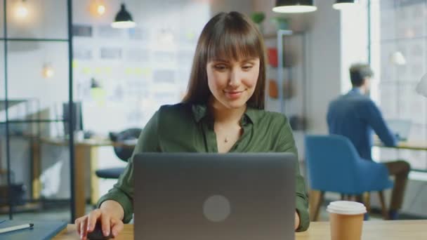 Giovane bella bruna lavora su un computer portatile in Cool Creative Agency in un Ufficio Loft. Ha un caffe 'da asporto e un quaderno sul tavolo. Lei sorride e ride. — Video Stock