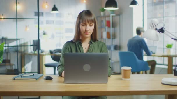 若い美しいブルネットはロフトオフィスのクールなクリエイティブエージェンシーのラップトップコンピュータで動作します。彼女はテイクアウェイコーヒーと彼女のテーブルのノートを持っています.彼女の同僚の仕事を背景に. — ストック動画