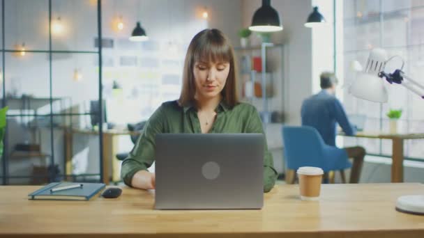 若い美しいブルネットはロフトオフィスのクールなクリエイティブエージェンシーのラップトップコンピュータで動作します。彼女はテイクアウェイコーヒーと彼女のテーブルのノートを持っています.彼女の同僚の仕事を背景に. — ストック動画