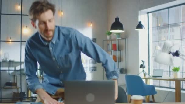 年轻英俊的男子在阁楼办公室的酷创意代理公司的笔记本电脑上工作。他桌上有一份外卖咖啡和一本笔记本。他穿着一件牛仔裤衬衫. — 图库视频影像