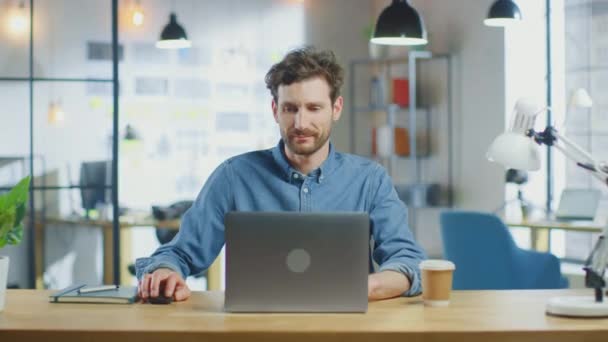 若いハンサムな男はロフトオフィスのクールなクリエイティブエージェンシーでラップトップコンピュータ上で動作します。テーブルの上にはテイクアウトコーヒーとノートがある。ジーンズのシャツを着ている。. — ストック動画
