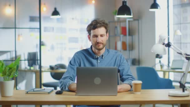 Joven hombre guapo trabaja en una computadora portátil en Cool Creative Agency en una oficina Loft. Tiene un café para llevar y un cuaderno sobre la mesa. Lleva una camisa vaquera.. — Vídeo de stock