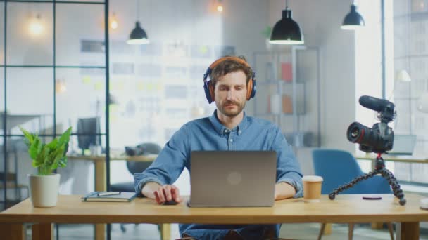 若いハンサムな男はロフトオフィスのクールなクリエイティブエージェンシーでラップトップコンピュータ上で動作します。テーブルの上にはテイクアウトコーヒーとノートがある。ジーンズのシャツとヘッドフォンを着て. — ストック動画