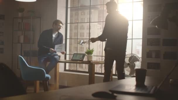 Twee jonge en knappe ontwerpers zitten op het bureau en bespreken Social Media App Interfaces op hun laptop. Ze werken in een Sunny Creative Office Loft met een groot raamzicht op Modern Urban City. — Stockvideo