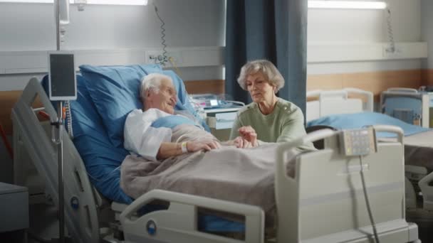 Hospital Ward Old Husband en la cama habla con su esposa visitando — Vídeos de Stock