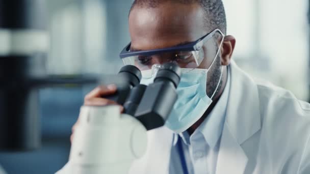 Scienziato maschio nero in maschera facciale utilizzando il microscopio in laboratorio — Video Stock