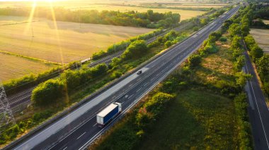 Aerial Drone Shot: Long Haul Semi Trucks Driving on the Busy Highway in the Rural Region of Italy. Agricultural Crop Fields and Hills in the Background clipart