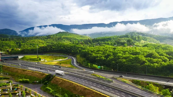 Aerial Drone Shot: Long Haul Semi Trucks Driving on the Busy Highway in the Rural Region of Italy. Beautiful Mountains with Forest and Clouds in the Background