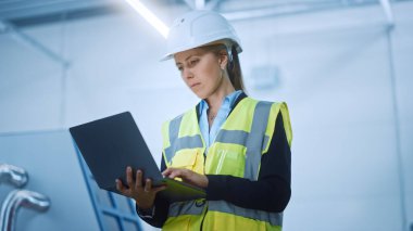 Yüksek Teknoloji Fabrikası: Güvenlikli Ceket Giyen Kendine Güvenen ve Profesyonel Kadın Mühendis ve Sıkı Şapka Tutma ve Bilgisayarda Çalışma. Modern Parlak Endüstriyel Tesis. Düşük Açı Vuruşu