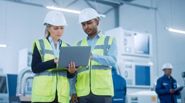 Güvenlik Yelekleri ve Sert Şapkalar Giyen Baş Mühendis ve Proje Yöneticisi Modern Fabrika 'da Laptop Kullan, Konuşuyor, Üretim Hattı İyimser. Sanayi Tesisi: Makineler üzerinde çalışan profesyoneller