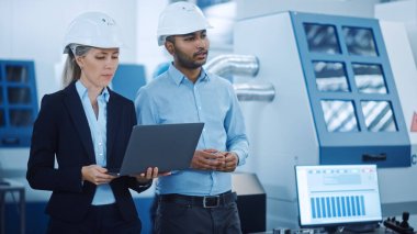 Güvenlik Yelekleri ve Sert Şapkalar Giyen Baş Mühendis ve Proje Yöneticisi Modern Fabrika 'da Laptop Kullan, Konuşuyor, Üretim Hattı İyimser. Sanayi Tesisi: Makineler üzerinde çalışan profesyoneller