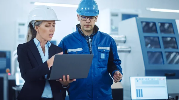 Gerente femenina e ingeniero de proyectos que usa cascos duros use el ordenador portátil en la fábrica industrial, hable, planifique la optimización de la productividad. Los trabajadores de la línea de producción operan maquinaria CNC, brazo robot del programa —  Fotos de Stock