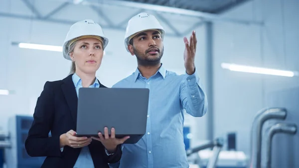 Ingénieur en chef féminin et gestionnaires de projets masculins se tenant au milieu de l'usine moderne, utiliser un ordinateur portable, planifier l'optimisation de la chaîne de production. Les professionnels travaillent sur des machines CNC industrielles — Photo