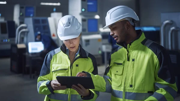 Ingeniero jefe y gerente de proyecto que usa chalecos de seguridad y sombreros duros, utilice la computadora digital de la tableta en la fábrica moderna, hablando, optimizando la línea de producción. Instalación industrial con maquinaria CNC —  Fotos de Stock