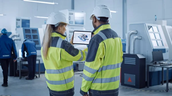 Industry 4.0 Factory: Chief Engineer e Project Supervisor in Safety Vests and Hard Hats, Talk, Use Digital Tablet Computer, Screen Shows 3D Concept of New Engine. Officina industriale con macchinari — Foto Stock