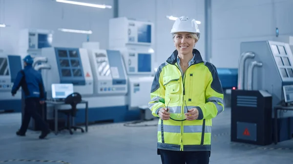 Όμορφη χαμογελαστή γυναίκα μηχανικός φορώντας αλεξίσφαιρο γιλέκο και Hardhat κατέχει γυαλιά ασφαλείας. Επαγγελματική γυναίκα που εργάζεται στο Σύγχρονο Εργοστάσιο Παραγωγής. Εγκατάσταση με CNC μηχανήματα και βραχίονα ρομπότ — Φωτογραφία Αρχείου