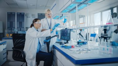 Modern Tıbbi Araştırma Laboratuvarı: Dijital Tablet kullanan iki kadın bilim adamı biyokimyasal örnekleri analiz ediyor. Tıp Bilimsel Laboratuvarı, Mikrobiyoloji Geliştirme. Gelişmiş Ekipman