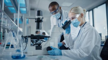 Modern Tıbbi Araştırma Laboratuvarı: Mikroskop kullanarak çalışan, Örnekleri analiz eden, konuşan yüz maskesi takan iki bilim adamı. Tıp ve Biyoteknoloji için Gelişmiş Bilimsel Laboratuvar.