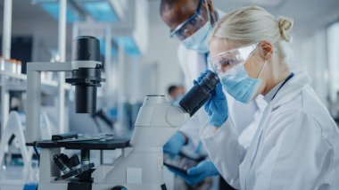 Modern Tıbbi Araştırma Laboratuvarı: Mikroskop kullanarak çalışan, Örnekleri analiz eden, konuşan yüz maskesi takan iki bilim adamı. Tıp ve Biyoteknoloji için Gelişmiş Bilimsel Laboratuvar.