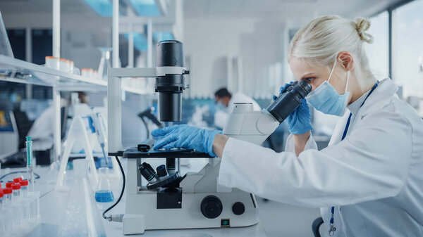 Modern Medical Research Laboratory: Portrait of Female Scientist Wearing Face Mask Looking Under Microscope, Analysing Test Samples. Advanced Scientific Lab for Medicine, Microbiology Development