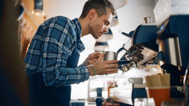 Checkered Shirt 'teki yakışıklı erkek barista bir kafede tek kullanımlık geri dönüşüm kupasında kapuçino yapıyor. Kadın Kasiyer, arka plandaki rahat bir kafede çalışıyor..