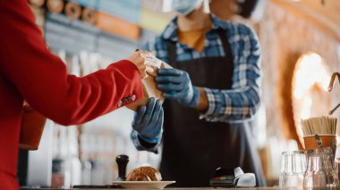 Kırmızı paltolu bir kadının, içinde hamur işi ve karton bardakla bir kafede kahvesini götürürken çekilmiş yakın çekimi. Konforlu Çatı Tarzı Kafe Tezgahının Arkasında Mutlu Çalışanlar Çalışıyor.