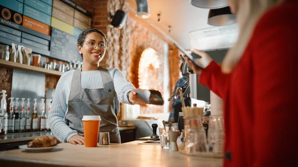 Joyful Multiethnic Diverse Woman Gives a Payment Terminal to Customer Using NFC Technology on Smartphone. Customer Uses Mobile to Pay for Take Away Latte and Croissant to a Barista in Coffee Shop.