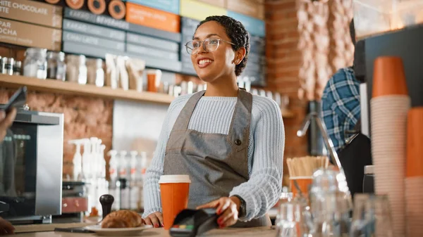Joyful Multiethnic Diverse Woman Gives a Payment Terminal to Customer Using NFC Technology on Smartphone. Customer Uses Mobile to Pay for Take Away Latte and Pastry to a Barista in Coffee Shop.