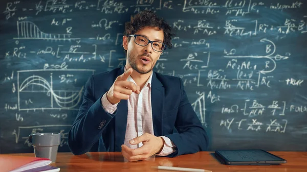 Online e-Education: Teacher Sitting at Desk, Explains Lesson to a Classroom, Behind Him Blackboard with Writing. e-Learning, Online Courses: Lecturer does Video Call, Explains Subjects to Students