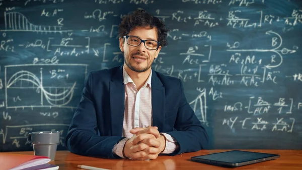 Online e-Education: Teacher Sitting at Desk, Explains Lesson to a Classroom, Behind Him Blackboard with Writing (engelsk). e-læring, nettkurs: Lærer gjør videokall, forklarer emner for elever stockbilde