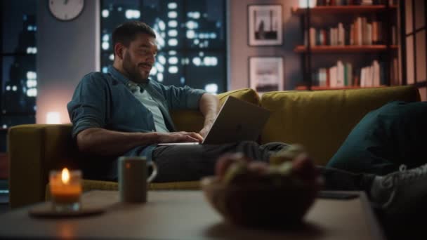 Чоловік з ноутбуком відпочиває у темній вітальні ввечері — стокове відео