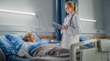 Hastane Koğuşu: Dost canlısı Doktor Yatakta Dinlenen Gülümseyen Yaşlı Kadın İçin Sağlık Bakımı Soruları Soruyor. Doktor tablet bilgisayar kullanıyor, kontrol ediyor, yaşlı kadınla tamamen iyileştikten sonra konuşuyor.