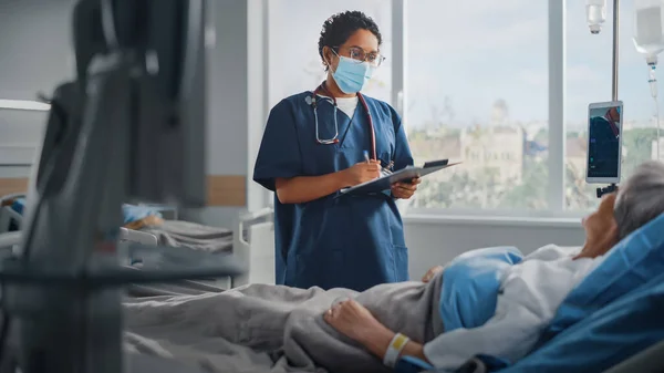 Salle d'hôpital : Une infirmière amicale portant un masque facial parle à une belle patiente âgée se reposant au lit, vérifie ses signes vitaux, rédige des informations. Infirmière en chef et vieille dame se rétablissant après Covid-19 — Photo