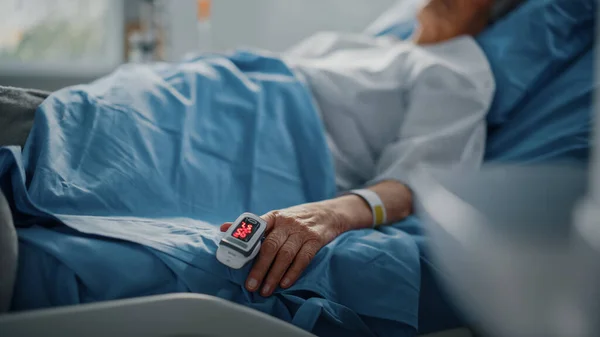病院病棟:シニア女性パルスを示す指心拍数モニターパルス酸素濃度計を備えたベッドで休んでください。彼女の壊れやすい手は毛布の上に休んで.手を中心に. — ストック写真