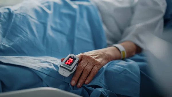 Hospital Ward: Senior Woman Descansando en una cama con Finger Heart Rate Monitor Oxímetro de pulso que muestra Pulso. Sus manos frágiles descansando sobre una manta. Concéntrate en la mano. — Foto de Stock