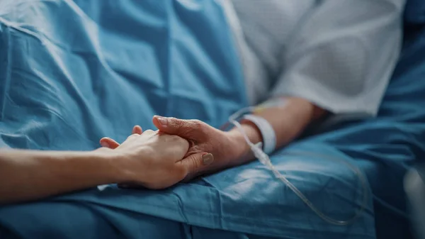 Hospital Ward: Senior Vrouw rust in een bed met Vinger Heart Rate Monitor, zorgzame familielid houdt haar fragiele hand, Ondersteuning en troost. Focus op de handen. — Stockfoto